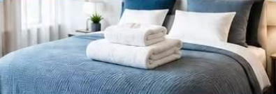 Neatly made bed with grey bedding, folded white towels and a bright window